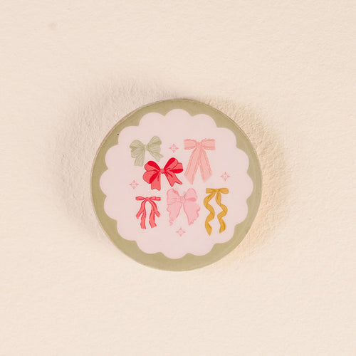 Store your Bow Affair Wood Enamel Coasters from The Darling Effect in style with this round tin, featuring a scalloped white label adorned with six colorful illustrated bows arranged in a circle on a light background.