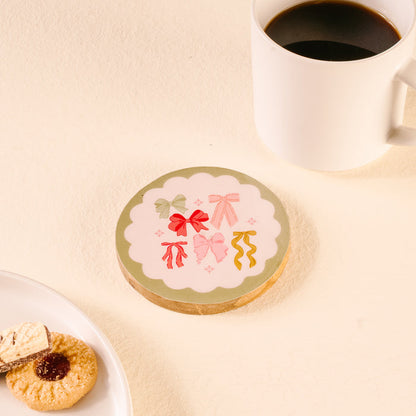 A white mug of black coffee rests on a table beside The Darling Effect's Bow Affair Wood Enamel Coaster. Nearby, assorted cookies and wafers sit on a plate, creating a cozy and inviting scene.