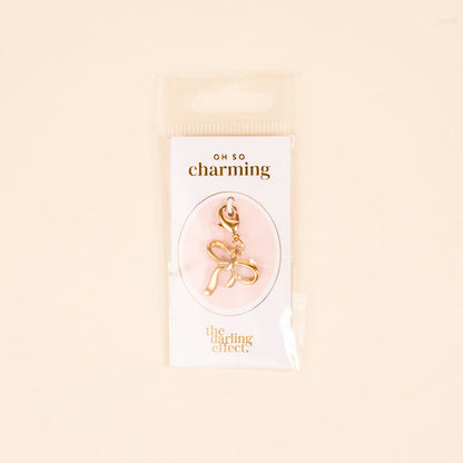 The Bow Jewelry Charm Gold by the-darling-effect is shown in clear packaging on a pink background. The text reads "OH SO charming" and "the darling effect," making it perfect for charm jewelry or cute necklaces.