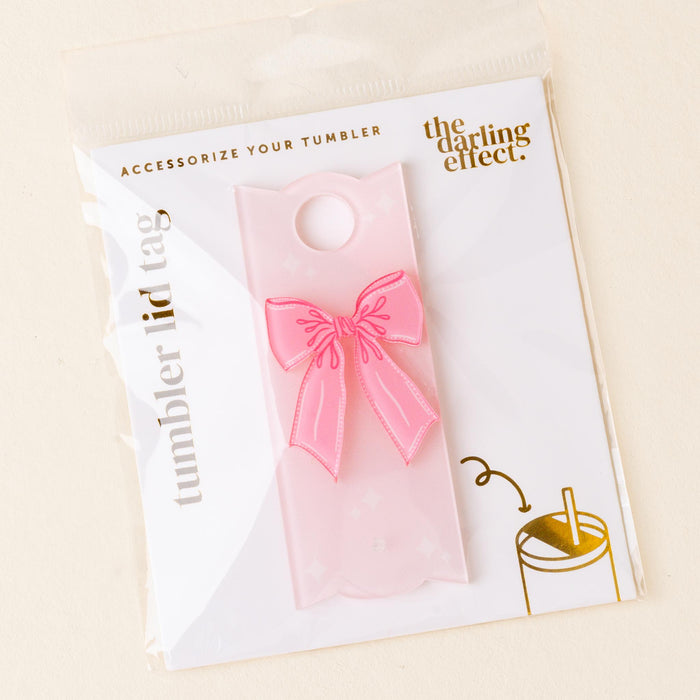 The Bow Pink Tumbler Lid Tag by the-darling-effect is shown in packaging. A pink translucent bow shape adds an adorable touch to your tumbler—perfect for personalizing your drinkware collection.