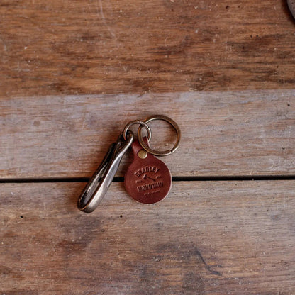 Forager Provisions LTD Bradley Mountain brass fish hook key fob with a branded leather tag on a rustic wooden surface.