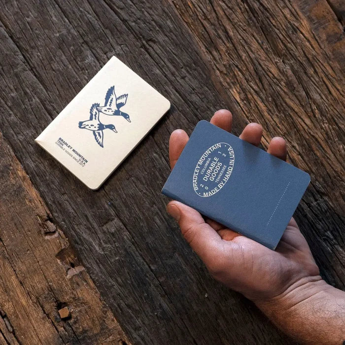 Hand holding blue Bradley Mountain Pocket Journal; another journal placed nearby, perfect for outdoor field note-taking.