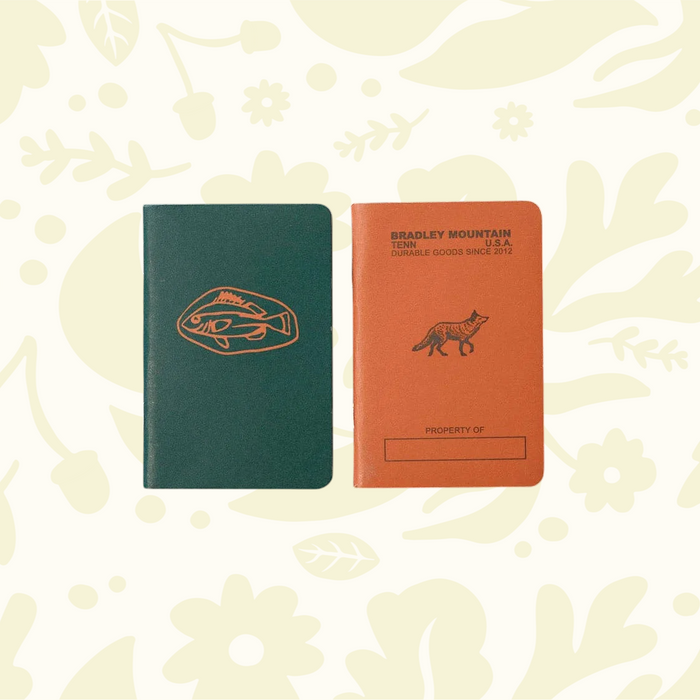 Forager Provisions LTD Bradley Mountain pocket journals in green and orange with fish and fox illustrations on wood.