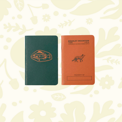 Forager Provisions LTD Bradley Mountain pocket journals in green and orange with fish and fox illustrations on wood.