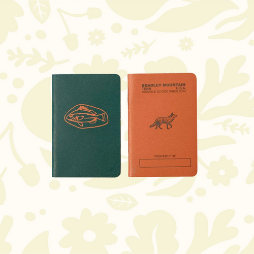 Forager Provisions LTD Bradley Mountain pocket journals in green and orange with fish and fox illustrations on wood.