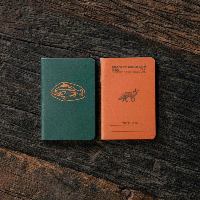 Forager Provisions LTD Bradley Mountain pocket journals in orange and green on a dark rustic wooden surface.