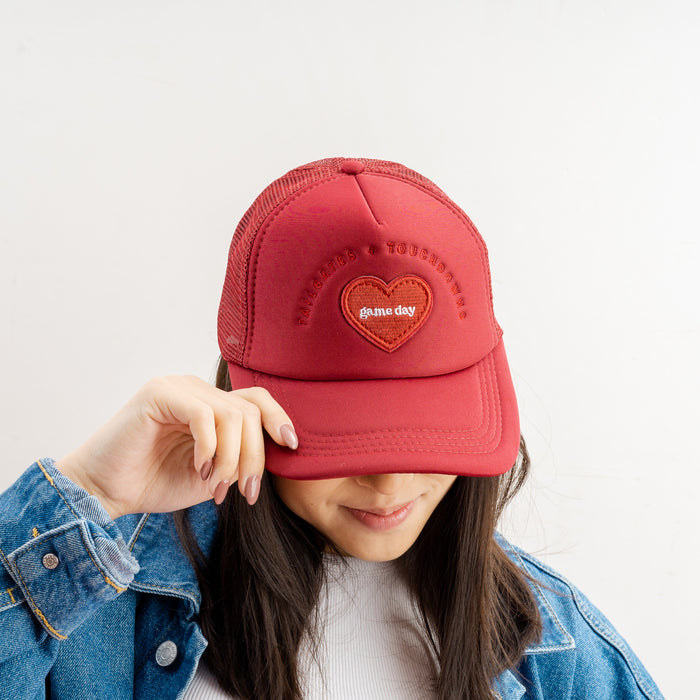 A person tilts down a Burgundy Game Day Trucker Hat by The Darling Effect, partially covering their face. They wear a blue denim jacket, adding subtle team spirit to their look.