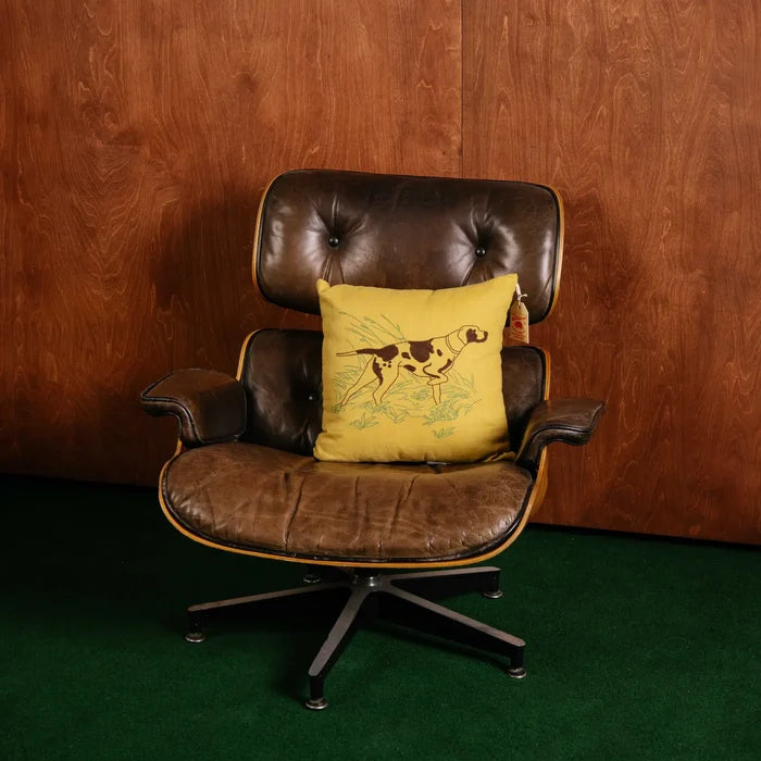 Vintage brown leather lounge chair with Camp Wandawega Pointer Pillow on green carpet, by retro wood-paneled wall.