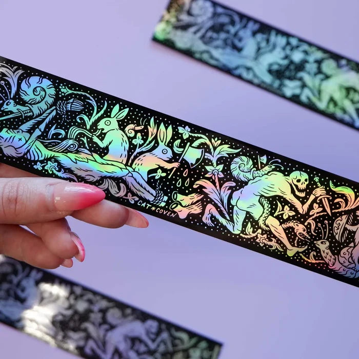 Hand holding Cat Coven Rabbits vs Skeletons holographic sticker with mystical art, light purple background, witchy aesthetic.