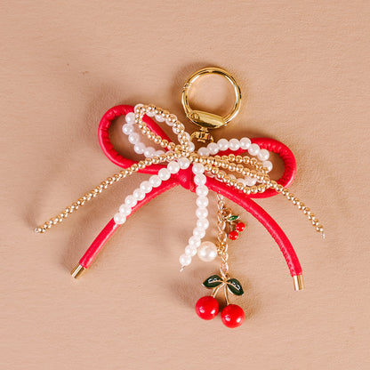 The Darling Effect's Cherry Bliss Layered Bows Bag Charm features gold keyring, layered red bows with pearls and gold beads, and a dangling cherry charm—an ideal statement or elegant accessory for your bag.