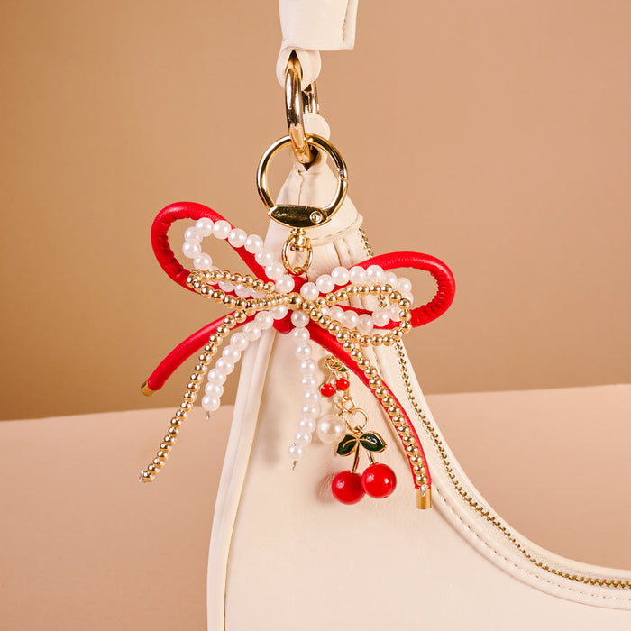 A close-up of a white handbag showcases The Darling Effect's Cherry Bliss Layered Bows Bag Charm, featuring red and pearl beaded bows, gold chains, and a cherry with leaf pendant on a gold clasp against a soft beige background.
