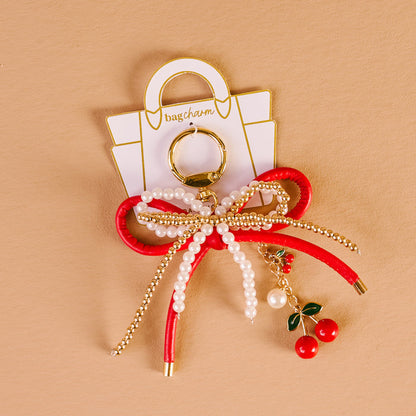The Darling Effect's Cherry Bliss Layered Bows Bag Charm features red and white ribbon, pearl beads, gold chains, and cherry ornaments on a handbag-shaped backing set against a beige background.