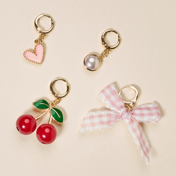 The Darling Effect’s Cherry on Top The Ultimate Charm Pack features four gold hoop charms—a pink heart, white pearl, red cherries with green leaves, and a pink-and-white gingham bow—perfect for scrunchie charm sets or accenting statement necklaces. Shown on beige.