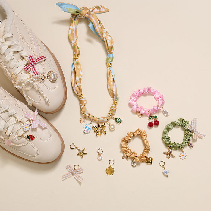 A flat lay of The Darling Effect's Cherry on Top The Ultimate Charm Pack shows white sneakers with shoe charms, a gold beaded scarf necklace with charms, and four scrunchie charm sets on a beige background, plus assorted individual charms and bows.