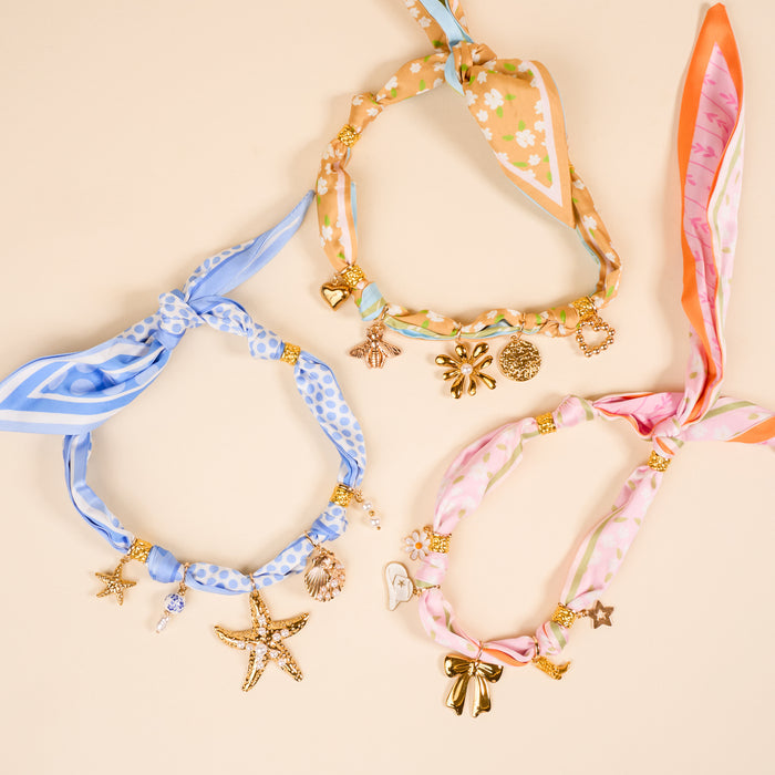 Cherry on Top The Ultimate Charm Pack by The Darling Effect features three vibrant silk scarves—blue, peach, and pink—with gold star, seashell, bow, and flower charms, inspired by scrunchie sets and displayed on a beige background.