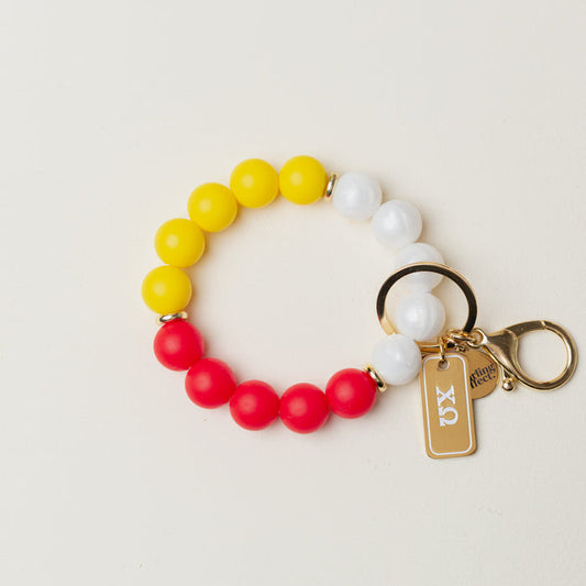 The Darling Effect Chi Omega Hands-Free Keychain Wristlet features yellow, white, and red silicone beads in a circle, gold key rings, a gold clasp, and a rectangular gold "JX" tag.