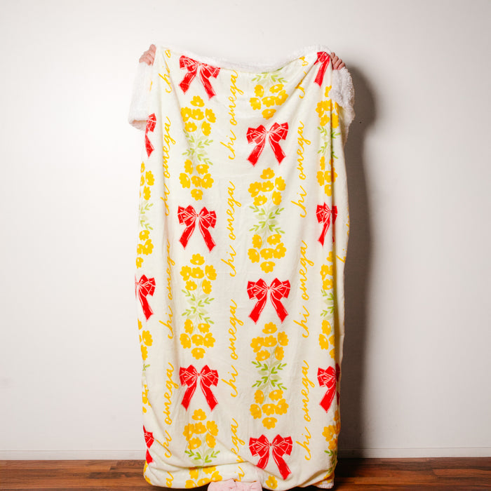 A person stands behind The Darling Effect's Chi Omega Plush Sherpa Blanket, featuring red bows, yellow flowers, and cursive script. Only their hands and feet show as the cozy blanket contrasts with a white wall and wooden floor.