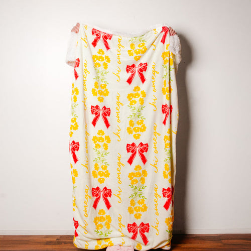A person stands behind The Darling Effect's Chi Omega Plush Sherpa Blanket, featuring red bows, yellow flowers, and cursive script. Only their hands and feet show as the cozy blanket contrasts with a white wall and wooden floor.