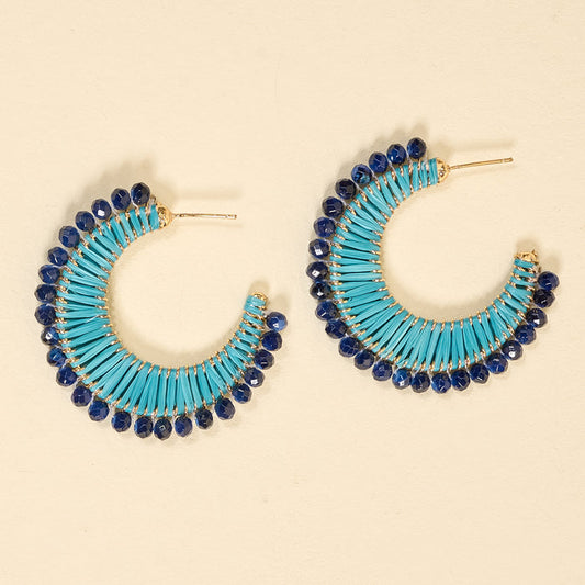 The Darling Effect's Coastal Blue It-Girl Earrings feature turquoise and navy raffia-wrapped hoops with gold studs and faceted dark blue crystals, ideal for daily wear—shown on a cream background.
