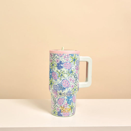 The Darling Effect’s Coastal Cove 20 oz Lifestyle Flip Straw Tumbler, featuring a tall insulated design with a large square handle and a colorful pink, blue, and green floral pattern, is displayed on a white surface against a beige background.