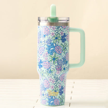 The Coastal Cove 40 oz Lifestyle Flip Straw Tumbler by the-darling-effect, featuring a large handle and leak-resistant flip straw lid, displays blue, pink, and green florals. It sits on a white surface with a beige backdrop—ideal for travel.