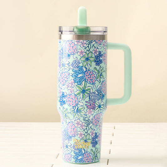 The Coastal Cove 40 oz Lifestyle Flip Straw Tumbler by the-darling-effect, featuring a large handle and leak-resistant flip straw lid, displays blue, pink, and green florals. It sits on a white surface with a beige backdrop—ideal for travel.