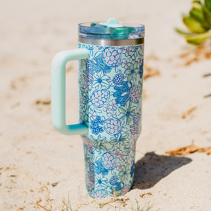 The-darling-effect Coastal Cove 40 oz Lifestyle Flip Straw Tumbler, featuring a blue handle and floral pattern with pink and blue flowers, rests on sandy ground in sunlight with a small green plant nearby.