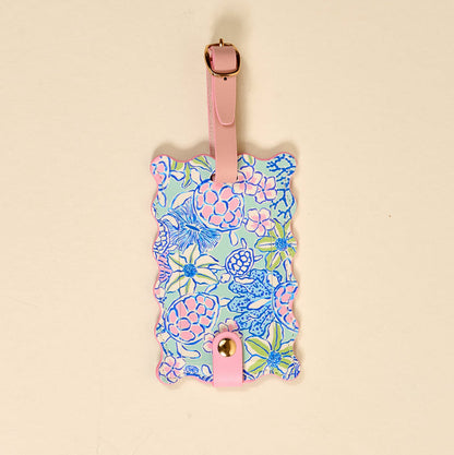 The Coastal Cove Jetsetter Luggage Tag is a rectangular tag with scalloped edges, a pastel blue, pink, and green floral print, privacy flap, and attaches to bags with a pink strap and gold buckle.