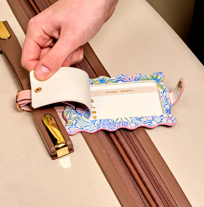 A hand lifts the privacy flap of the Coastal Cove Jetsetter Luggage Tag on a beige suitcase, revealing the name and contact info card inside this stylish travel accessory.