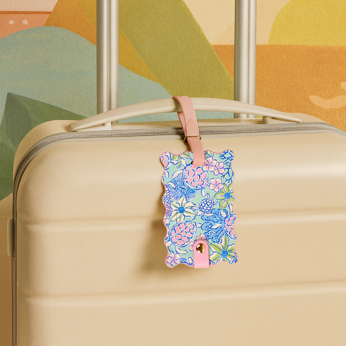 A beige suitcase with the Coastal Cove Jetsetter Luggage Tag—an essential travel accessory with pink, blue, and green hues—attached to the handle, set against a vibrant abstract wall mural.