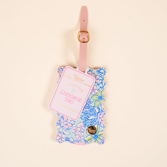The Darling Effect’s Coastal Cove Jetsetter Luggage Tag features a floral print in pink, blue, and green on a beige background, with a pink strap, gold buckle, privacy flap, and “Jetsetter Luggage Tag” text.