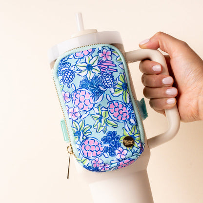 A hand holds a large white tumbler with a handle, featuring the-darling-effect’s bestselling Coastal Cove Tumbler Fanny Pack—a small, water-resistant zippered pouch with a blue, pink, and green floral and berry pattern.