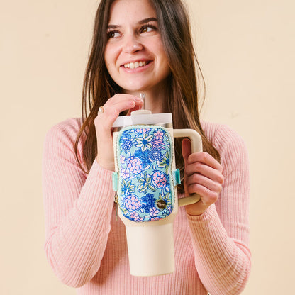 A smiling woman in a pink sweater drinks from a beige tumbler featuring the darling-effect's bestselling Coastal Cove Tumbler Fanny Pack with a floral pattern, set against a plain beige background.