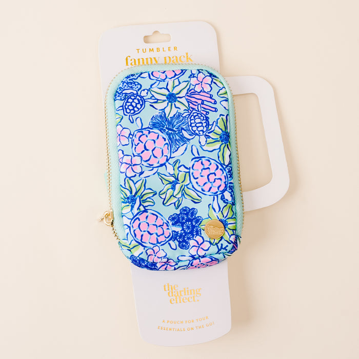 A colorful, floral Coastal Cove Tumbler Fanny Pack from the-darling-effect sits on a cream background with branded packaging and a gold heart-shaped zipper pull—this bestselling, water-resistant accessory is perfect for any outing.