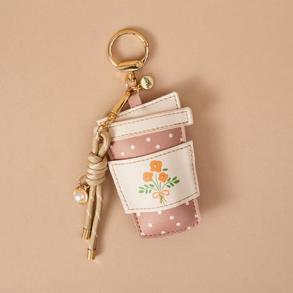 The Darling Effect’s Coffee Run Vegan Leather Bag Charm—shaped like a pink polka-dotted coffee cup with a white sleeve adorned with orange flowers, a gold key ring, and a knotted cord with a pearl charm—makes the perfect playful accessory for your bag. Beautifully displayed on a beige background, this charming piece is an ideal choice for anyone seeking fun Vegan Leather Bag Charms.