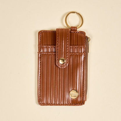The Darling Effect’s Cognac Ripple Ridge Keychain Wallet features vertical ribbing, a gold keyring, snap closure, zippered coin holder, and small round gold emblem on the bottom right, shown on a beige background.