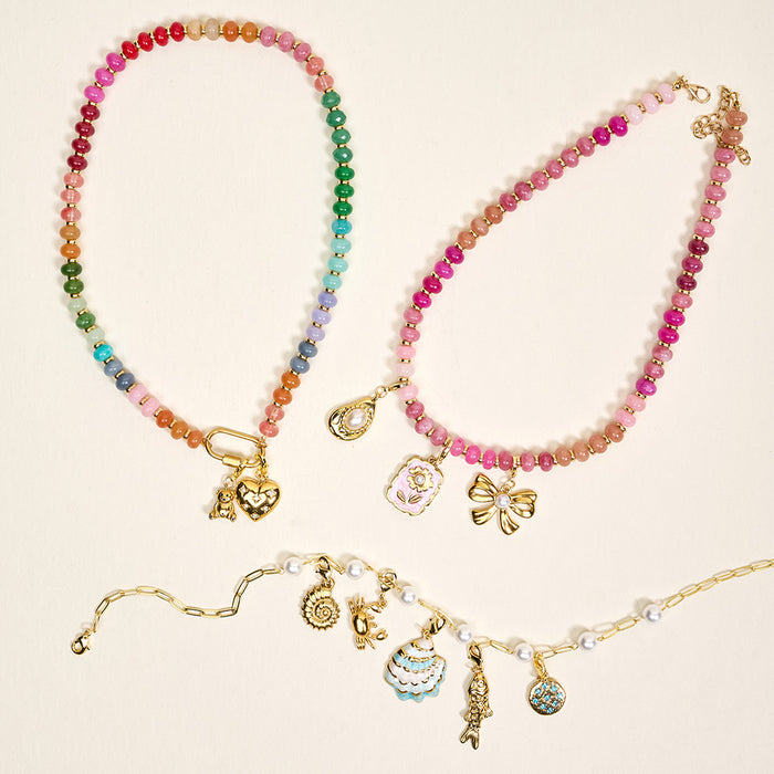 Three beaded necklaces with gold clasps from the-darling-effect feature dangling charms, including the Compass Jewelry Charm Gold, hearts, flowers, and bows—a playful charm bar jewelry display on a light background.