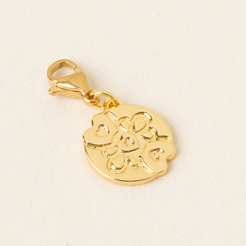 The Compassionate Jewelry Trait Charm Gold by the-darling-effect, featuring an engraved floral design and lobster clasp, rests on a light cream background—an elegant symbol of confidence and strength.