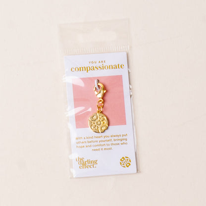 The-darling-effect Compassionate Jewelry Trait Charm Gold comes with a clasp, packaged on a card stating "You are compassionate" with the brand’s logo below, all inside a clear plastic sleeve.