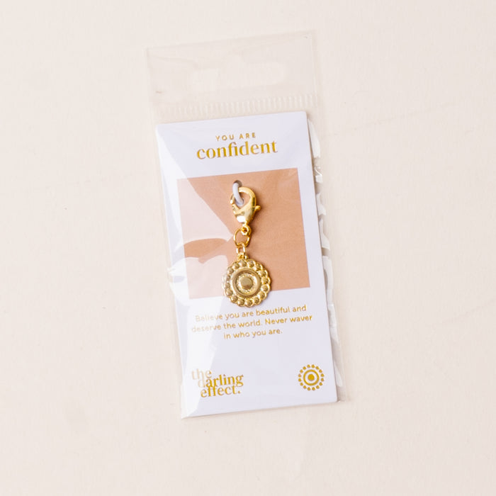 The-darling-effect Confident Jewelry Trait Charm Gold, featuring a decorative circular design, is attached to a card reading, "You are confident. Believe you are beautiful and deserve the world. Never waver in who you are.