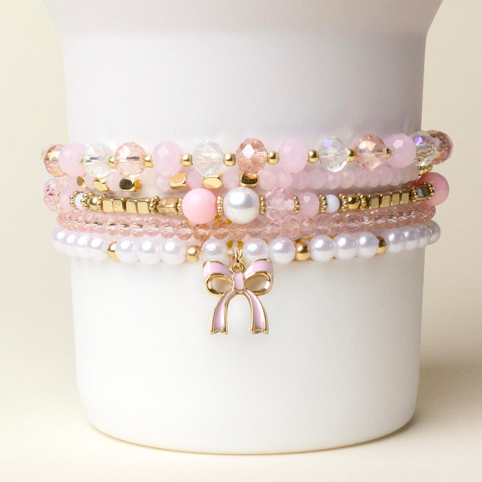 The-darling-effect Coquette Charm Tumbler Bracelet Stack features pink, white, and gold beaded bracelets with a gold bow charm—perfect as chic tumbler accessories or a stylish bracelet stack.