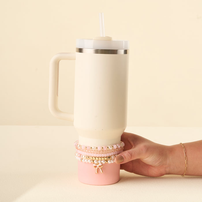 A hand wearing a gold bracelet holds a 40 oz cream and pink tumbler with a handle and straw, featuring the-darling-effect's Coquette Charm Tumbler Bracelet Stack on the base for extra flair.