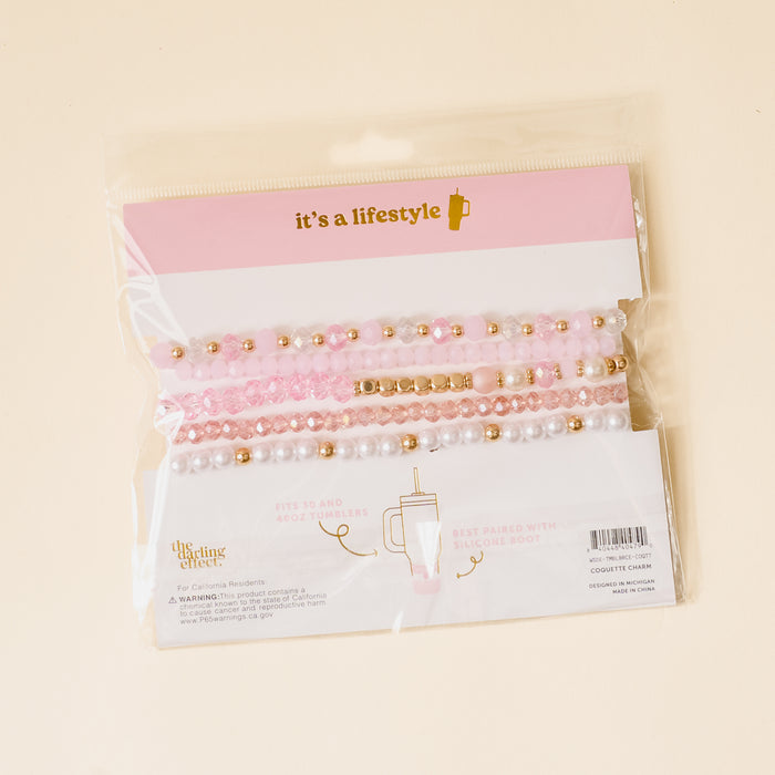 The-darling-effect’s Coquette Charm Tumbler Bracelet Stack features pink, white, and gold beaded charms in clear packaging with a pink and white label—perfect accessories for your 40 oz tumbler.