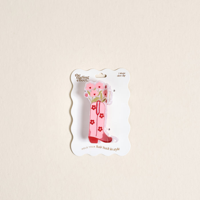 The Darling Effect’s Cowgirl Boot Shape Up Claw Clip is a pink acrylic claw clip shaped like a cowboy boot with floral design, displayed on a wavy white backing card.
