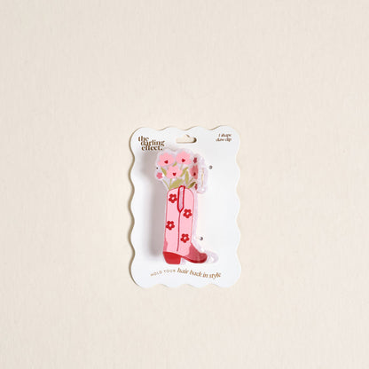 The Darling Effect’s Cowgirl Boot Shape Up Claw Clip is a pink acrylic claw clip shaped like a cowboy boot with floral design, displayed on a wavy white backing card.