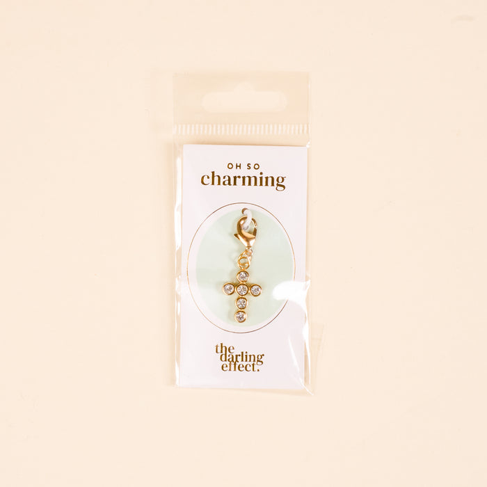 The Cross Jewelry Charm Gold by the-darling-effect features sparkling rhinestones and is attached to an OH SO charming card, packaged in clear plastic against a light beige background—perfect for charm jewelry.