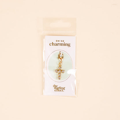 The Cross Jewelry Charm Gold by the-darling-effect features sparkling rhinestones and is attached to an OH SO charming card, packaged in clear plastic against a light beige background—perfect for charm jewelry.