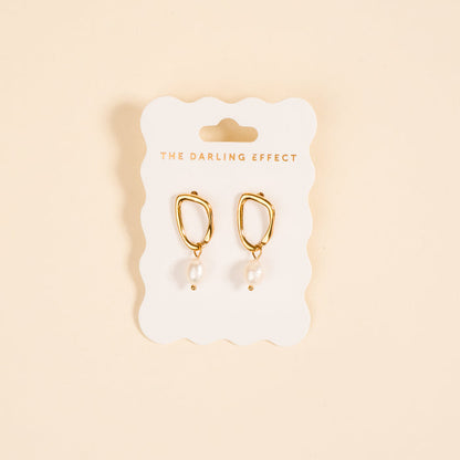 The Dainty Drift Standout Earrings by The Darling Effect feature gold-tone shine and dangling white pearls, displayed on a branded card against a beige background—ideal for adding elegance to any special occasion.
