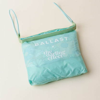 The Daisy Craze Aqua Ballast Beach Pillow by The Darling Effect features a folded teal design with florals, white branding, light blue straps, and a mint green accent—perfect for stylish outings and available as a limited-edition collaboration.