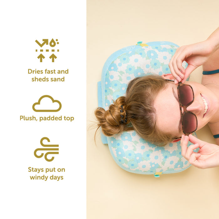 A woman in sunglasses relaxes on The Darling Effect’s Daisy Craze Aqua Ballast Beach Pillow. Icons highlight its fast-drying, sand-shedding plush top and wind-resistant design. Shop this limited-edition collab now!.